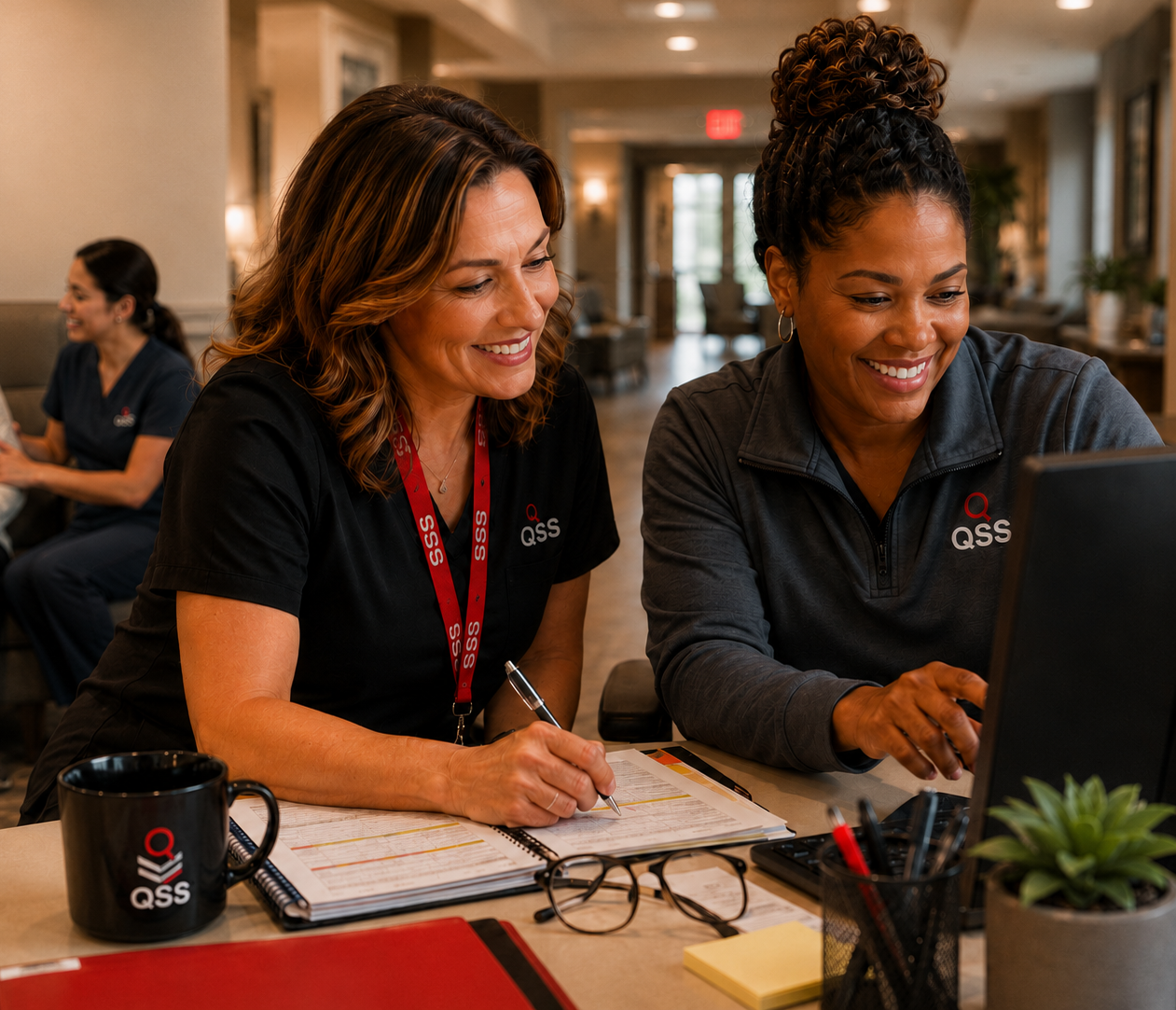 Two QSS team members collaborating at a desk, reviewing documents and a monitor together, with a resident visible in the background